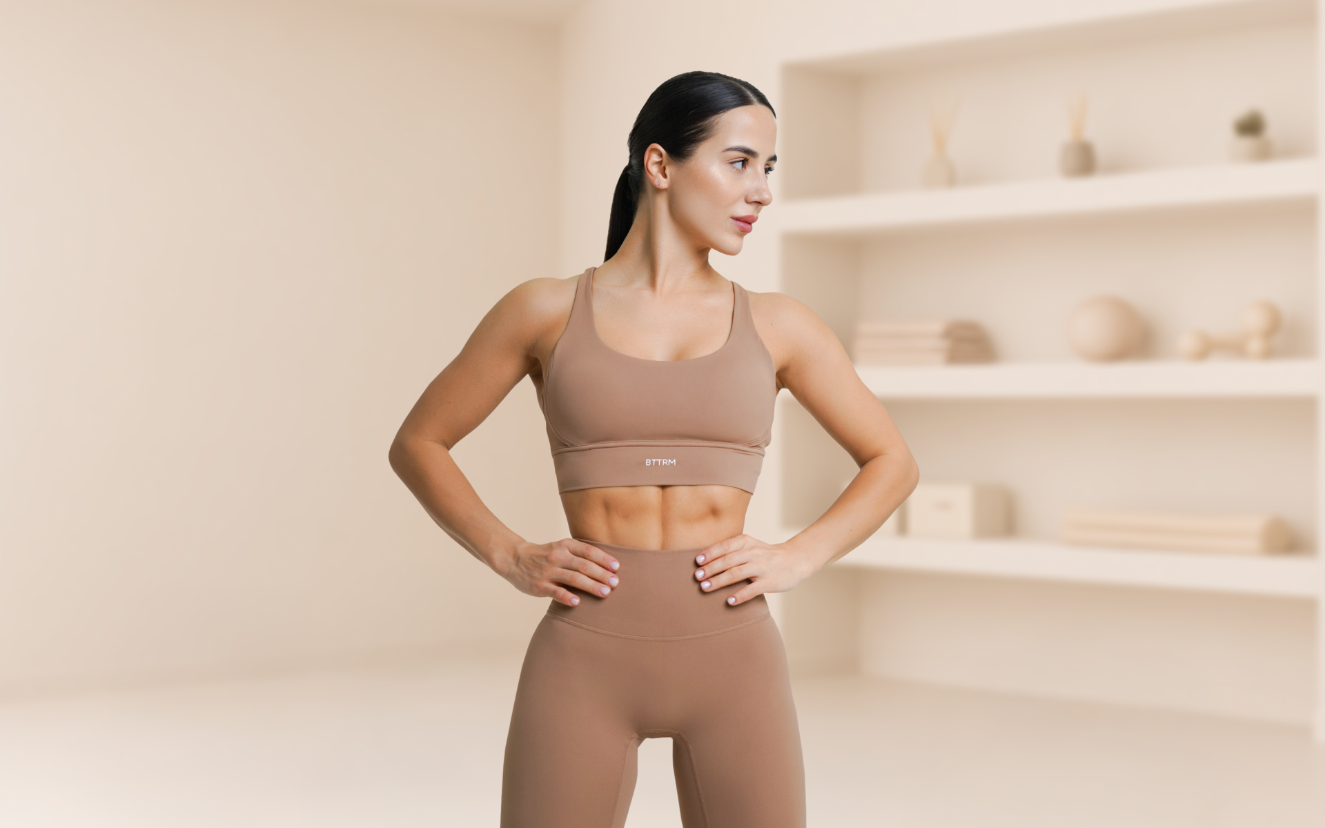 Lessie Fitness in a beige activewear set stands with hands on hips in a bright studio, looking to the side.